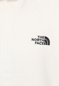 Abbigliamento in pile bianco con zip e logo ricamato nero di The North Face nella parte sinistra del petto.
