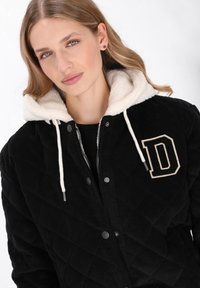 Fekete kordbársony dzseki steppelt textúrával, krémszínű gyapjúbéléses kapucnival, húzózsinórral és krémszínű "D" betűs díszítéssel a mellkason.