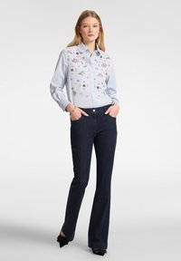 Camicia azzurra chiara con ricami floreali sul petto, abbinata a jeans a zampa neri e scarpe con tacco nere.