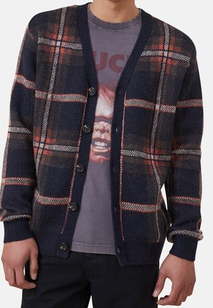 Mann trägt eine marineblaue, rot-weiße karierte Strickjacke mit Knöpfen über einem ausgewaschenen T-Shirt mit einem Gesicht und Buchstaben.