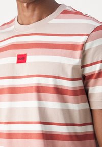 Stripet t-shirt med korte ærmer i nuancer af pink og rød med hvide detaljer. Har en rød rektangulær lap med teksten "HUGO" på brystet.