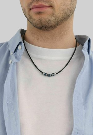 Mann mit leichtem Stoppelbart, der ein blaues offenes Hemd über einem weißen T-Shirt trägt, mit einer schwarzen Kordelhalskette mit silbernen und blauen Perlen.