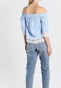 Blouse à rayures bleu et blanc, épaules dénudées, avec bordure en dentelle, associée à un jean bleu clair. La blouse présente une coupe décontractée et un col élastique.