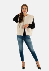 Gilet senza maniche beige chiaro, in lana strutturata, camicia nera a maniche lunghe sotto, jeans blue fitted e scarpe nere con tacco alto.
