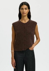 Brązowy bezrękawnik z okrągłym dekoltem, teksturowana dzianina, z czarnymi guzikami na przodzie. W zestawieniu z ciemnoniebieskimi dżinsami.