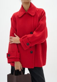 Rode wollen jas met een brede kraag, grote zwarte knopen, oversized mouwen met manchetten. Model houdt een bruine handtas vast en draagt donkere broeken.