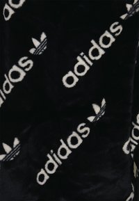 Černá imitace kožešiny s opakujícím se bílým logem "adidas" a motivem trojlístku, poskytující měkký, strukturovaný vzhled.