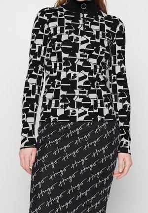 Femme portant un haut à manches longues à motif géométrique noir et blanc avec col zippé et une jupe noire avec un texte blanc en cursive répété "Hugo".