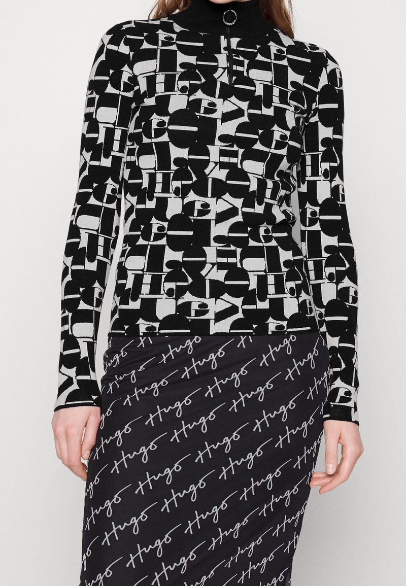 Femme portant un haut à manches longues à motif géométrique noir et blanc avec col zippé et une jupe noire avec un texte blanc en cursive répété "Hugo".