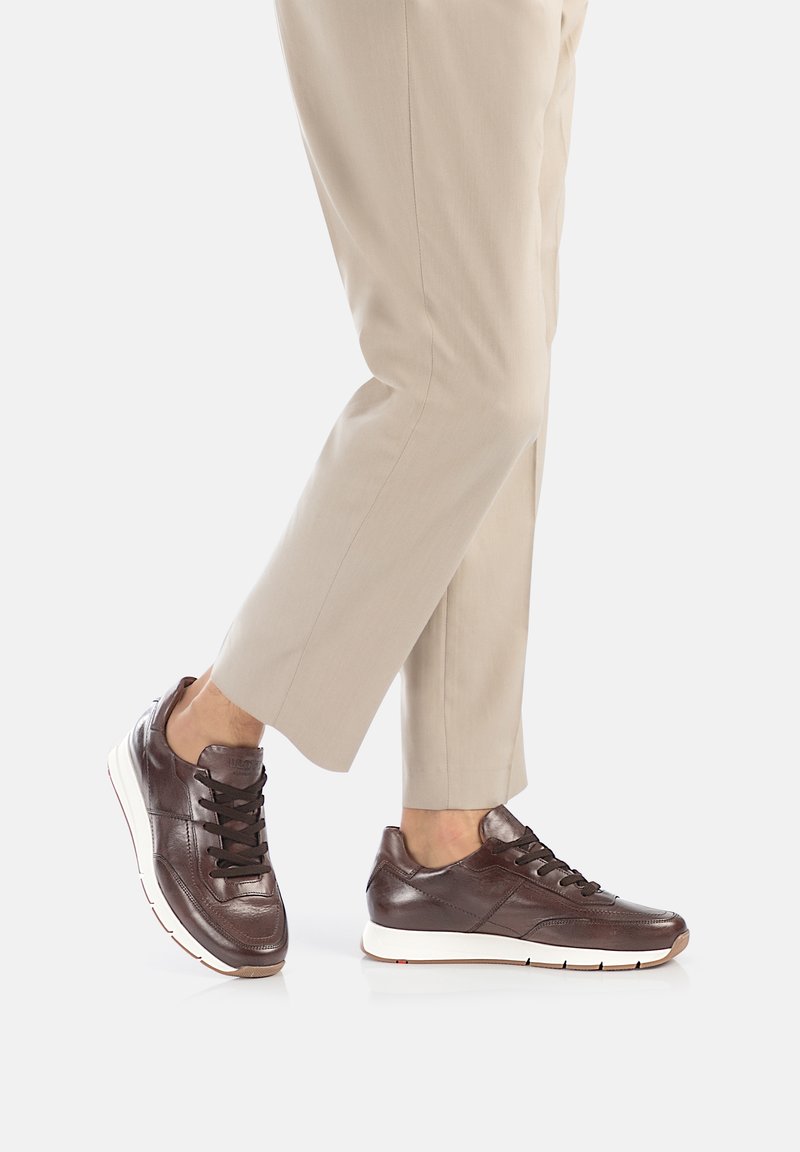 Sneakers in pelle marrone con punta rotonda, lacci neri, colletto imbottito e suola in gomma bianca. Indossati con pantaloni beige su misura.