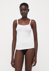 Witte camisole met dunne spaghettibandjes, gemaakt van gladde katoenstof. Heeft een vierkante halslijn en een getailleerde pasvorm. Zwarte bikini slip zichtbaar.