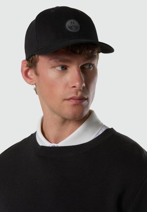 Giovane uomo che indossa un cappellino da baseball nero con un logo circolare North-Sails, un maglione nero e una camicia bianca con colletto, che guarda verso destra.