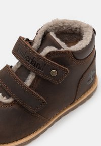 Bottine en cuir marron avec deux bandes Velcro, doublure en polaire, détails cousus et un logo embossé sur le côté. Semelle en caoutchouc plate.