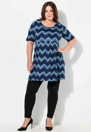 Kleid in Marineblau mit einem Zickzackmuster in verschiedenen Blautönen, kurzen Ärmeln und einem gerafften Bund, kombiniert mit schwarzen Leggings und High Heels.