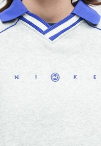 Άτομο φορώντας γκρι και μπλε πουκάμισο με γιακά με μπλε και λευκές ρίγες σε σχήμ α V και κεντημένο λογότυπο "NIKE" στο στήθος.