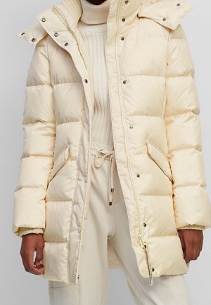 Beige gesteppte Steppjacke mit Druckknöpfen und Reißverschluss, getragen über einem cremefarbenen, gerippten Rollkragenpullover und passenden Jogginghosen mit Kordelzug.