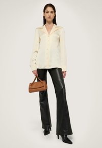 Lys gul skjorte med krave, parret med sorte læder flared bukser. Modellen holder en brun håndtaske.