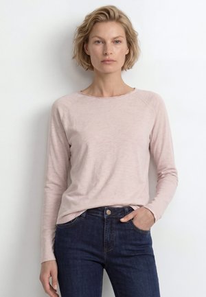 Femeie blondă stă pe un fundal alb, purtând un tricou cu mânecă lungă roz deschis și blugi bleumarin, cu o mână în buzunar.
