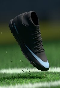 Schwarzer Fußballschuh mit hohem Schnitt, mit einem blauen Nike-Swoosh. Strukturierte Oberfläche und sichtbare Schnürsenkel, leicht vom Boden abgehoben.