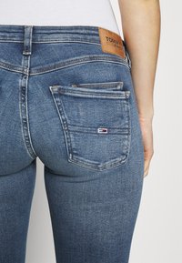 Blue denim jeans featuring a mid-rise waist, two back pockets, and a leather logo patch on the waistband. Smooth texture and fitted design.