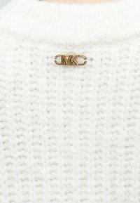 Λευκό πουλόβερ πλεκτό με υφή και χρυσό λογότυπο MK καρφίτσα στην πλάτη. Το ύφασμα φαίνεται μαλακό και άνετο.