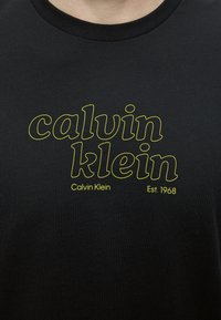 Μαύρο μπλουζάκι με κίτρινο περίγραμμα κειμένου "calvin klein" και μικρότερο κείμενο "Calvin Klein" και "Est. 1968" κάτω από τα γράμματα με περίγραμμα.