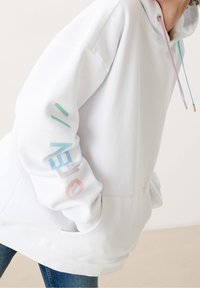 Sweat à capuche blanc avec une poche avant, présentant des graphismes en texte pastel sur la manche et des cordons de serrage colorés en rose et bleu. Texture douce.