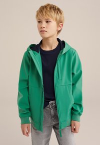 Giacca verde con cappuccio, chiusura con zip, tasche a marsupio e polsini a coste. Indossata sopra una camicia blu scuro e abbinata a jeans grigi.