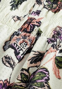 Tela con un patrón floral en base crema, que presenta un intrincado detalle en rosa, púrpura y verde sobre un algodón texturizado, fruncida en las costuras.