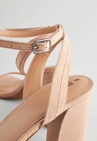 Beige högklackade sandaler med en mockaliknande yta, med korsade remmar, en metallspänne och en kraftig klack.