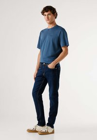 Modrá krátka tričko s krátkym rukávom v kombinácii s tmavými džínami; tričko má jednoduchý dizajn, džínsy majú svetlé švy a klasický strih. Pohodlné tenisky.