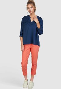 Marinebluse mit V-Ausschnitt und gerafften Details, kombiniert mit korallfarbenen cropped Hosen. Das Modell trägt weiße Sneaker. Einfaches, lässiges Design.