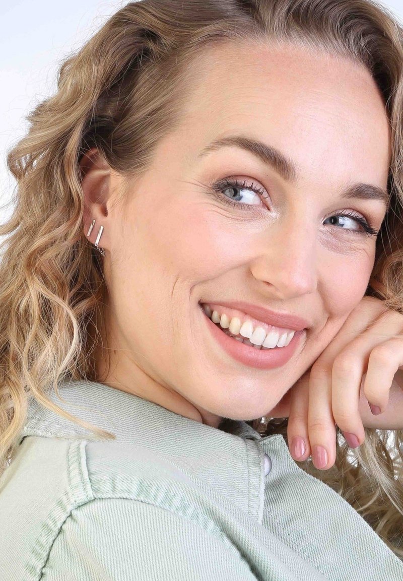 Boucles d'oreilles linéaires en argent présentées sur un modèle aux cheveux ondulés, sur fond clair. Le modèle porte une veste vert clair.