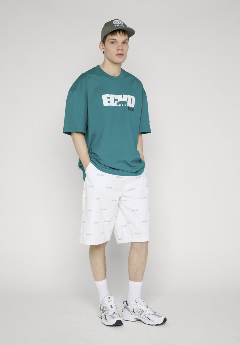 Jeune homme debout, les mains dans les poches, portant un t-shirt oversize turquoise Ecko United, un short blanc, des baskets blanches, des chaussettes, et une casquette verte, sur fond uni.