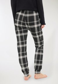 Persona a piedi nudi che indossa pantaloni a quadri bianchi e neri e una maglietta nera a maniche lunghe, in posizione eretta su uno sfondo grigio chiaro, vista da dietro.