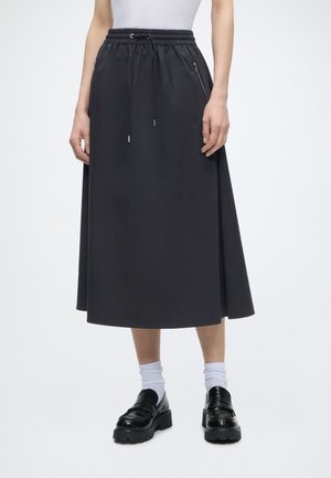 Moteris dėvi juodą midi sijoną su elastinga juosmens juostele ir raišteliu, derintą su baltais kojinėmis ir juodais stambiais loafers batais.