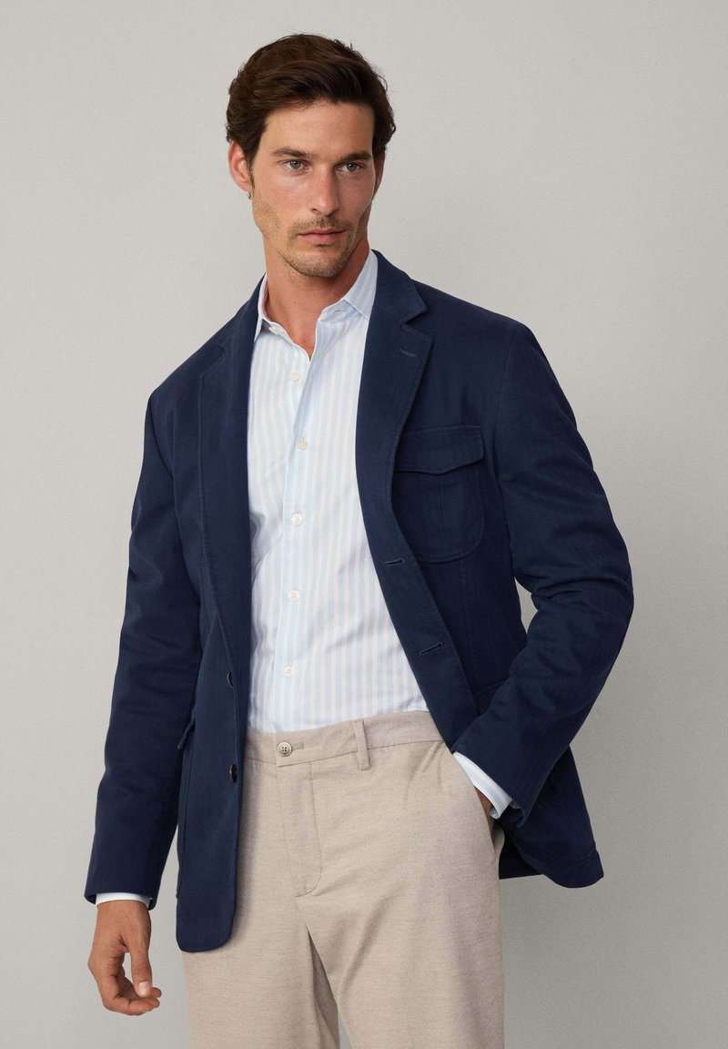 Blazer de algodón marino con un solo bolsillo en el pecho, llevado sobre una camisa a rayas azul claro, combinado con pantalones beige, mostrando un ajuste entallado.