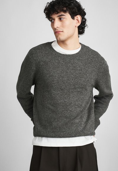 Pull en tricot gris foncé avec un col rond et des manches longues, superposé sur une chemise blanche, présentant un tissu texturé.