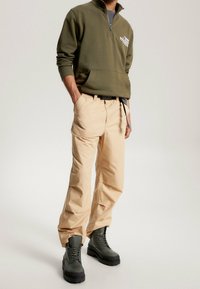 Olivově zelená mikina na zip s přední kapsou, spárovaná s béžovými volnými kalhotami a tmavozelenými šněrovacími botami s robustní podrážkou.