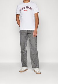 Bílá tričko s potiskem "GUESS JEANS 1981" v bordó barvě. Šedé džíny s texturovaným povrchem, v kombinaci s bílými teniskami s béžovými podrážkami.