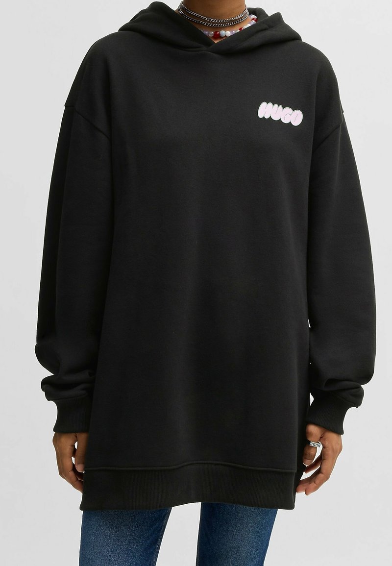 Personne portant un sweat à capuche noir oversize avec le logo blanc « HUGO » sur la poitrine, associé à un jean bleu et des colliers de perles en couches.
