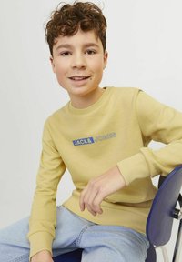 Felpa gialla con polsini a coste, con un logo rettangolare blu "JACK & JONES". Indossata con jeans azzurri, seduto su una sedia blu.