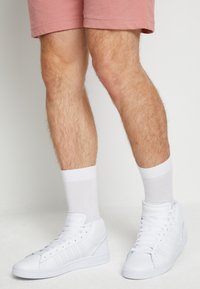 Persona vestida con zapatillas altas blancas, calcetines blancos, y pantalones cortos rosas, de pie sobre un suelo blanco contra una pared blanca.