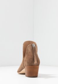 Bruine suede enkellaars met een ronde neus, een ritssluiting aan de achterkant en een gestapelde hak. De binnenkant is lichtbruin van kleur. Glad van textuur.