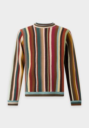 Homme debout portant un pull à rayures verticales multicolores, un jean large bleu clair et des baskets beiges sur un fond blanc uni.