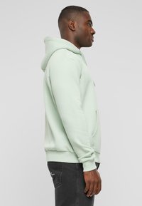 Felpa verde menta con tasca frontale, polsini e orlo a coste. Indossata con jeans neri, mostrando una vista laterale del fit rilassato e del design.