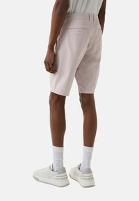 Une personne portant un short rose clair, des chaussettes blanches et des baskets blanches, se tenant contre un fond uni, vue de dos et à partir de la taille.