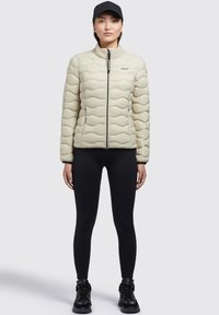 Jachetă puffer bej deschis cu design cu cuvete în formă de valuri, fermoar frontal, guler înalt, purtată peste colanți negri și cizme negre, cu o șapcă neagră.