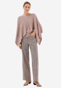 Svetle růžový pletený pončo s volným střihem, doplněné hnědými kostkovanými kalhotami širokého střihu. Modelka má na sobě béžové ankle boots na nízkém podpatku.