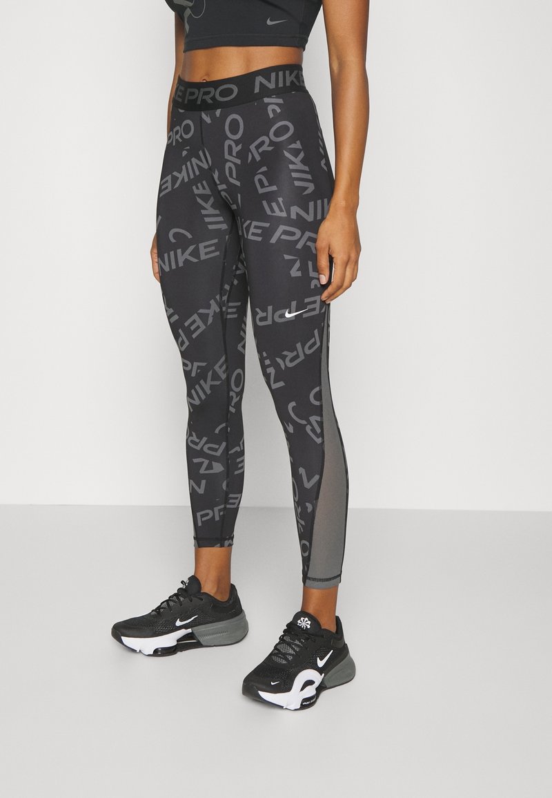 Leggings atléticos negros que presentan un patrón de logotipos grises repetidos, una cinturilla alta y paneles de malla a lo largo de los lados. Llevados con zapatillas negras.